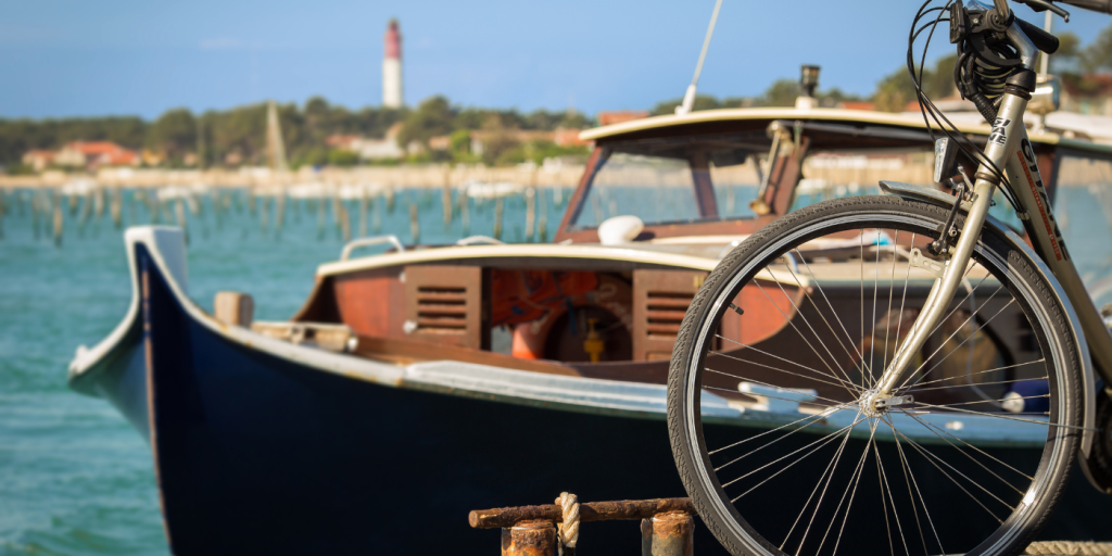 L'avant d'un vélo au premier plan, une pinasse au second et le Cap Ferret et son phrase en fond, floutés.
