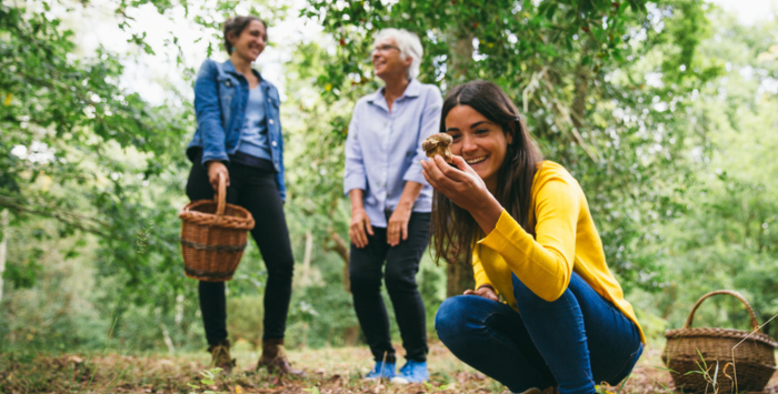 1 jeune femme au premier plan qui tient un cèpe dans ses mains dans une forêt d'Arès. À l'arrière plan, une jeune femme tenant un panier en osier, et une dame plus agée qui sourit